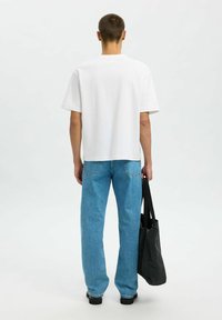 White short-sleeve t-shirt, relaxed fit, paired with light blue straight-leg jeans. Individual holds a dark tote bag, facing away.