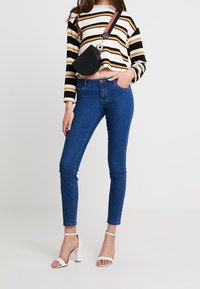 Suéter a rayas de ganchillo en negro, blanco y amarillo; jeans ajustados de cintura alta en azul; bolso de hombro negro; sandalias de tacón blancas.