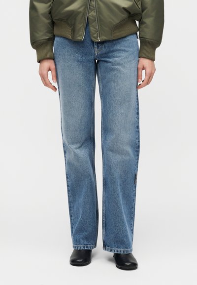 Άτομο που φοράει ανοιχτό μπλε τζιν με ίσια γραμμή, μαύρα παπούτσια και πράσινο χακί bomber μπουφάν, στέκεται μπροστά σε λευκό φόντο.