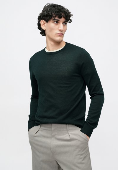 Dark green knit sweater with a textured diamond pattern, crew neckline, and ribbed cuffs, paired with light gray trousers.