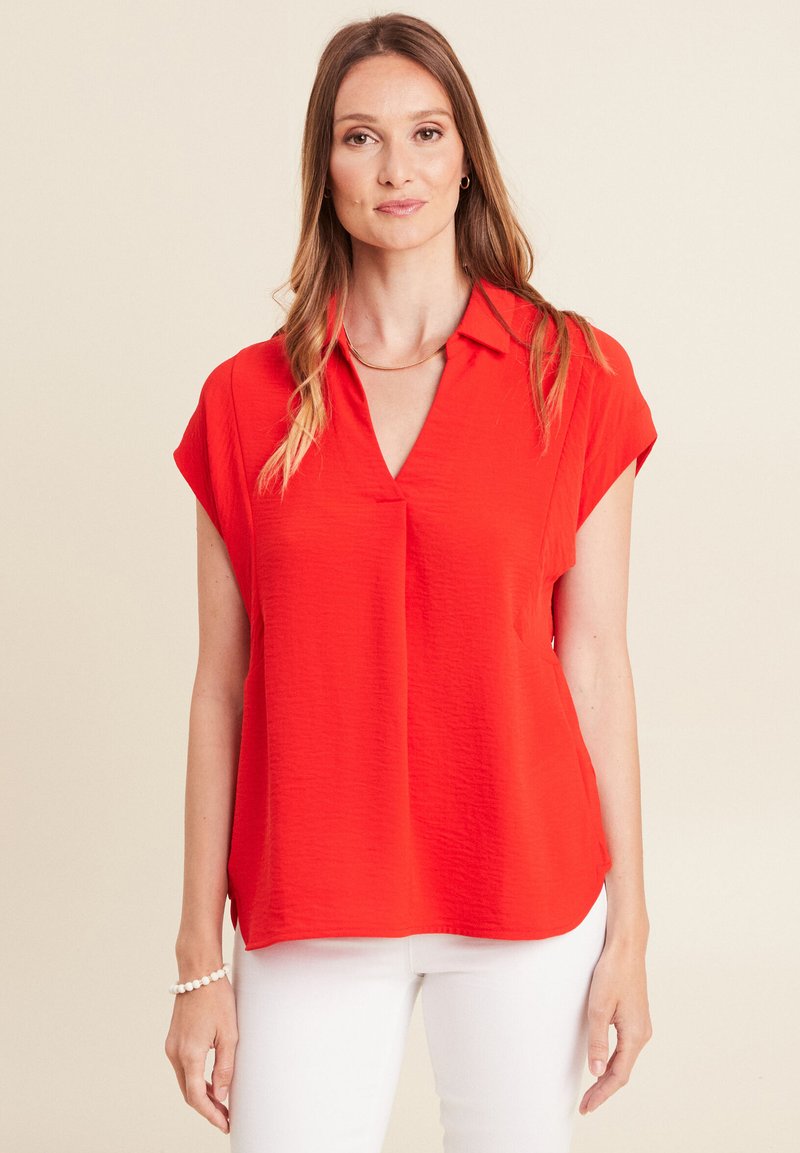 Blusa rossa a maniche corte con scollo a V e un tessuto leggermente testurizzato, vestibilità morbida e spacchi laterali, indossata su pantaloni bianchi.