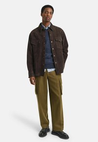 Braune Wildlederjacke mit Knopfleiste und zwei Brusttaschen, über einem blauen Pullover getragen, kombiniert mit grünen Cargo-Hosen und schwarzen Schuhen.