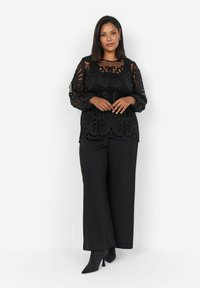 Top lungo a maniche di pizzo nero con intagli floreali, abbinato a pantaloni neri a gamba larga e stivaletti neri scintillanti.