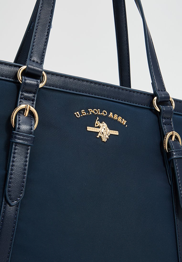 Sac fourre-tout bleu marine en tissu lisse avec deux poignées, présentant des détails en doré et un logo en relief à l'avant.