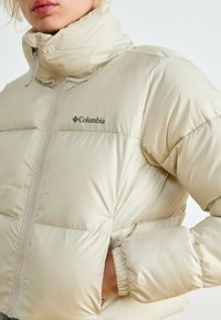 Veste matelassée beige avec un col montant, fermeture éclair et poignets élastiques. Comprend un petit logo Columbia noir sur la poitrine.