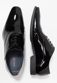 Une paire de chaussures habillées en cuir verni noir avec des lacets, une chaussure montrant une vue de dessus et l'autre une vue de côté sur fond blanc.