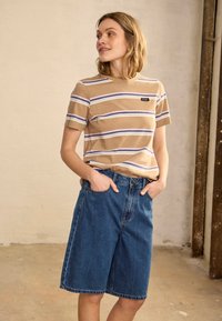 Femme aux cheveux blonds portant un t-shirt beige rayé et un short en jean bleu ample, debout les mains dans les poches dans une pièce simple.