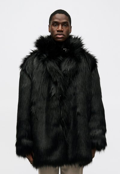 Mladý muž v černém kožichu z umělé kožešiny s objemným límcem, stojící na pozadí prosté bílé zdi.