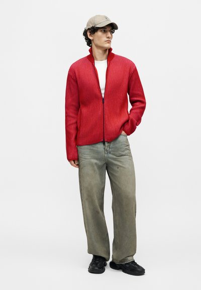 Νεαρός άνδρας που φοράει μπεζ καπέλο, κόκκινο ριπ πουλόβερ με φερμουάρ πάνω από λευκό πουκάμισο, φαρδιά ξεθωριασμένα γκρι τζιν και μαύρα αθλητικά παπούτσια, στέκεται με τα χέρια στις τσέπες.