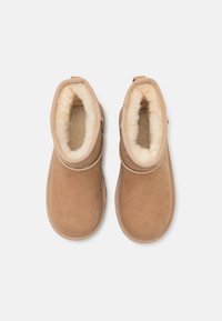 Botas de tobillo de ante color marrón claro con forro de piel de oveja crema, puntera redonda y costuras decorativas a lo largo del borde superior. Suela de goma para mayor tracción.