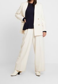 Nainen pukeutuneena kermaan vivahtaviin leveälahkeisiin housuihin, kermaan kaksiriviseen bleiseriin, mustaan ribbineuleeseen ja kermaan värisiin nilkkureihin, seisomassa käsi lantiolla.