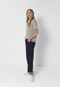 Žena s dlouhými blond vlasy, oblečená v béžovém svetru, tmavých srolovaných džínách a bílých teniskách, stojící s rukama v kapsách na bílém pozadí.