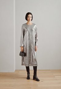 Srebrna sukienka wyszywana cekinami z długimi rękawami, okrągłym dekoltem i luźnym fasonem, w połączeniu z czarnymi botkami sięgającymi do kolan i małą czarną torebką.