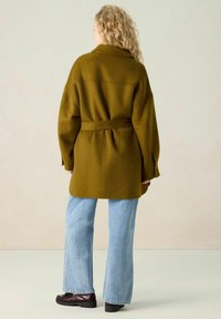 Manteau vert olive au design oversized, ceinturé à la taille, avec des manches larges, associé à un jean flare bleu clair et des chaussures noires épaisses.