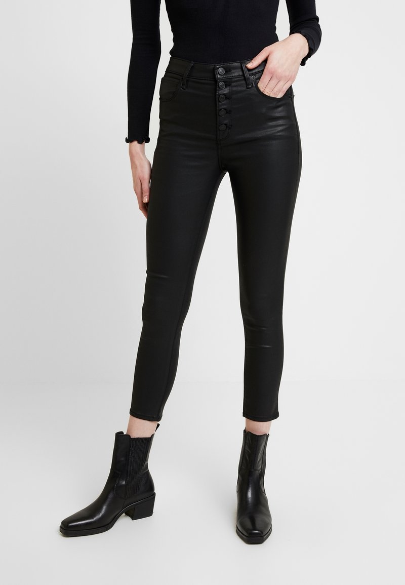 Černé kalhoty z umělé kůže s vysokým pasem, tvarovaným střihem, pěticí knoflíků vpředu a délka ke kotníkům, doplněné černými kotníkovými botami na podpatku.