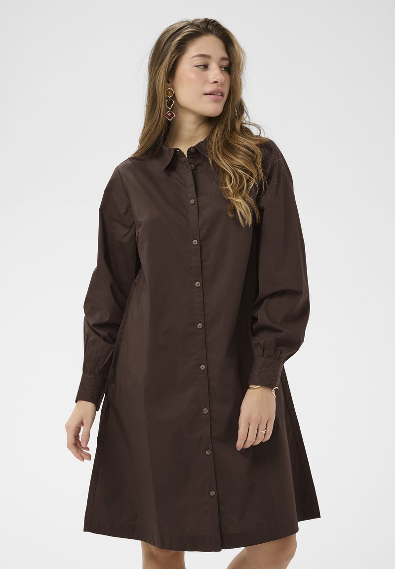 Femme portant une robe boutonnée marron foncé à manches longues avec des poignets bouffants, un bracelet en or, une bague et des boucles d'oreilles géométriques pendantes, regardant sur le côté.