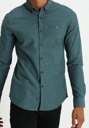 Hombre que lleva una camisa ajustada de manga larga y cierre de botones en color verde azulado con un pequeño logo bordado en amarillo en el pecho izquierdo.