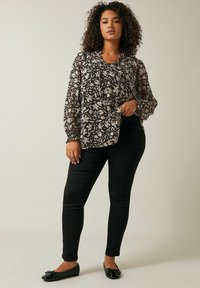 Blusa nera trasparente a fantasia floreale con maniche lunghe e arricciate, abbinata a pantaloni neri aderenti e scarpe basse scure.