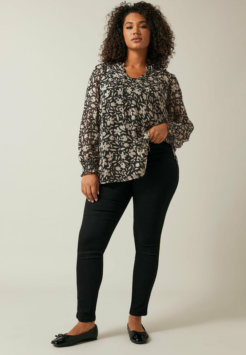 Blusa nera trasparente a fantasia floreale con maniche lunghe e arricciate, abbinata a pantaloni neri aderenti e scarpe basse scure.