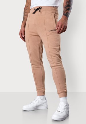 Helle beige Jogginghosen mit einem Kordelzugbund, Seitentaschen und vertikalen Nähten, kombiniert mit weißen Sneakers für einen Kontrast.