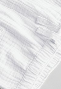 Tejido de muselina blanca con un patrón texturizado y arrugado. Presenta bordes fruncidos y dos lazos de tela para un cierre seguro.