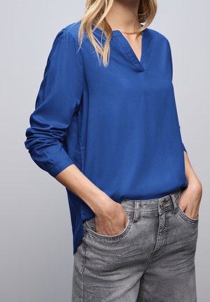 Vrouw met een blauw overhemd met lange mouwen en een V-hals, gedeeltelijk ingestopt in grijs gewassen spijkerbroek, met handen in de zakken.