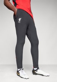 Muž v černých texturovaných tréninkových nohavicích Liverpool FC a bílých fotbalových kopačkách se zlatým logem, stojící s rukama v kapsách.