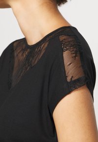 Svart kortärmad topp med genomskinliga spetssatser på axlarna, rundad halsringning och mjuk tygtextur.