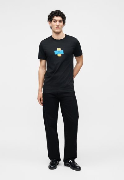 Jeune homme aux cheveux bouclés portant un t-shirt noir avec un motif coloré en forme de bande, un pantalon noir et des chaussures noires, debout devant un fond uni.