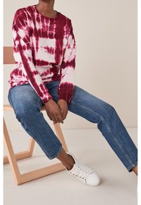 Maglietta a maniche lunghe con effetto tie-dye in bordeaux e bianco, con scollatura a girocollo, abbinata a jeans blu e sneakers bianche. Seduto su una sedia di legno.