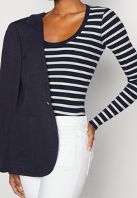 Marinblå blazer med en enkel ficka som hänger över en vit och marinblå randig långärmad topp, i kombination med vita jeans.