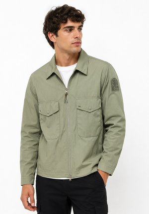 Homme portant une veste zippée vert clair avec poches à rabats et un écusson sur le bras, associée à un pantalon noir, regardant sur le côté.