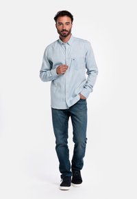 Lichtblauw overhemdje, lange mouwen, met een enkele borstzak, gedragen met donkere jeans en zwarte schoenen. De stof lijkt zacht en lichtgewicht te zijn.
