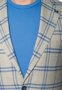 Jasno beżowy żakiet w niebieską kratę, zestawiony z jednolitym niebieskim koszulą. Żakiet ma klapę szpiczastą i jedno zapięcie na guzik.