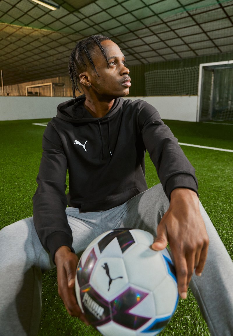 Zwarte hoodie met een wit logo, grijze joggers, zittend op kunstgras, met een zwart-witte voetbal met iriserende accenten.