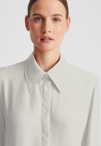 Weiße Bluse mit langen Ärmeln aus einem glatten Stoff. Verfügt über einen klassischen Kragen und eine durchgehende Knopfleiste. Steppnähte an den Kragenkanten.