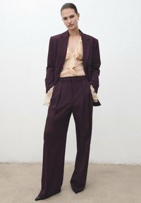 Traje entallado en burdeos oscuro con pantalones de pierna ancha y una chaqueta ajustada sobre una blusa nude con volantes en el escote.