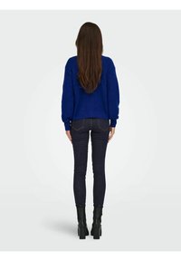 Modro pleten pulover z rebrastimi manšetami in spodnjim robom, kombiniran s temnimi ozkimi kavbojkami in črnimi gležnjarji. Vidni so dolgi ravni lasje.