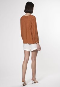 Blusa ambra con tessuto testurizzato, maniche lunghe e polsini con bottoni. Abbinata a pantaloni corti bianchi arrotolati e sandali con tacco bianchi.