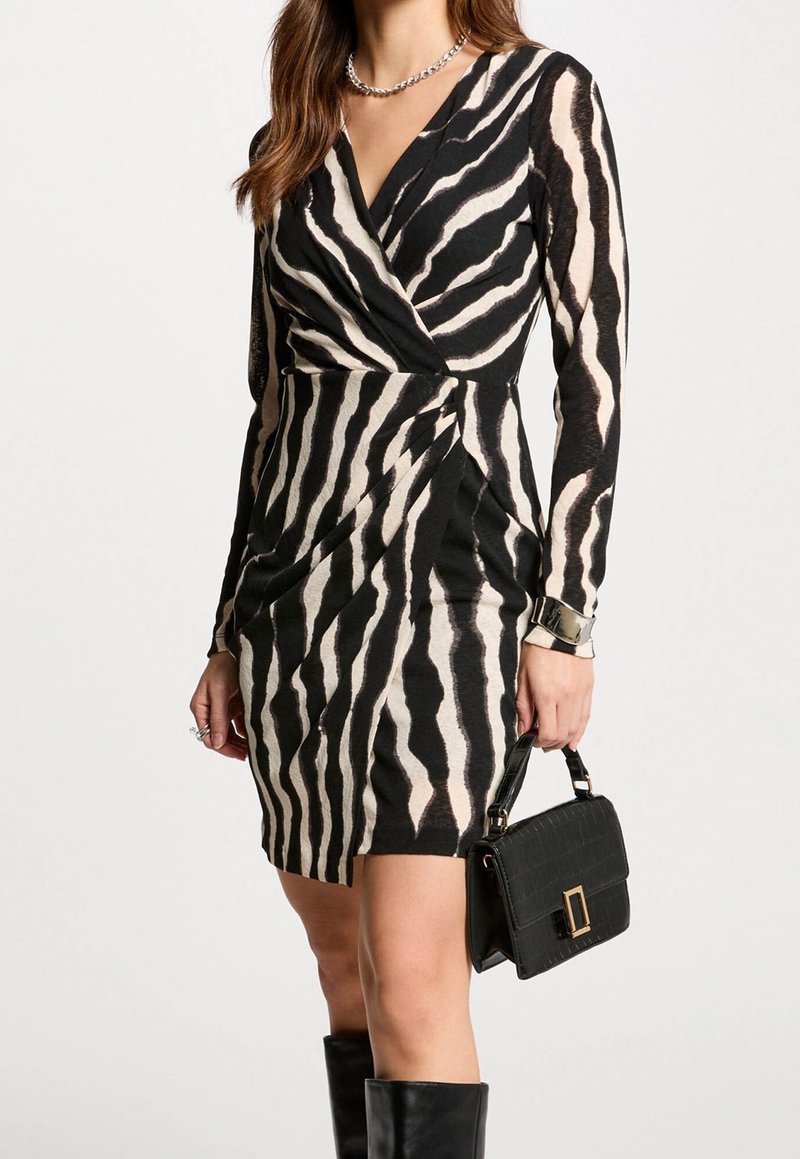 Femme portant une robe portefeuille imprimé zèbre noir et blanc, tenant un sac à main noir, avec des bottes noires montant jusqu'aux genoux et un collier en argent.