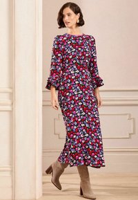 Rochie cu imprimeu floral, cu mâneci lungi și volane, decolteu rotund și o siluetă fluidă în culori vibrante pe un fundal întunecat.