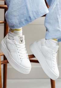 Weiße Leder-Sneaker mit strukturierten Akzenten, flachen Schnürsenkeln und einer erhöhten Gummisohle. Mit Markenetikett auf der Zunge und seitlichen Details.