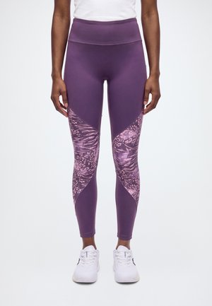 Le mannequin porte un legging violet taille haute avec des empiècements roses à motifs sur le bas des jambes, un haut blanc et des baskets blanches.
