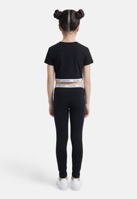 Παιδί με κοτσίδες διπλό κότσο, φοράει μαύρο crop top με κοντά μανίκια και κολάν με λευκή ζώνη Calvin Klein και λευκά αθλητικά παπούτσια, από πίσω.