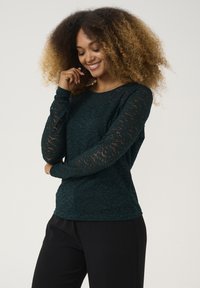Mörk teal långärmad topp med blommig spetsstruktur, solid kropp och genomskinliga sp sleeves. I kombination med svarta byxor har outfiten en avslappnad passform.