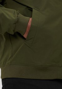 Oliven grøn jakke med en glat tekstur, ribbede manchetter og kant. Har en sidelomme og et strømlinet design.