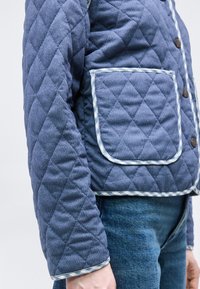 Μπλε μπουφάν με ραφές σε σχήμα διαμαντιού, μεγάλο εμπρόσθιο τσέπη, κουμπιά και καρό τελείωμα, φορεμένο πάνω από μπλε τζιν παντελόνι.