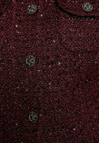 Tessuto bordeaux con texture e paillettes, con bottoni metallici e colletto leggermente rialzato. Due tasche frontali visibili.