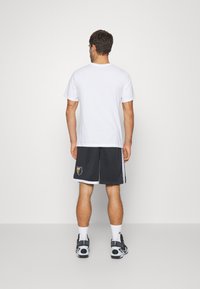 T-shirt blanc en coton à manches courtes, associé à un short de sport noir avec un logo et des bandes sur le côté. Porte des baskets noires.