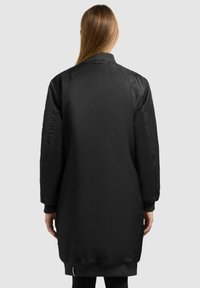 Giubbotto bomber nero, in materiale nylon liscio, lungo fino al ginocchio, con polsini e orlo a coste, spalle imbottite, dotato di uno spacco sul retro per facilitare i movimenti.